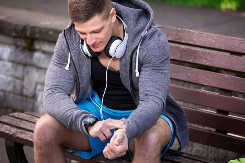 young man sitting on the bench in park and checking out his pulse rate - post on How To Lower Resting Heart Rate