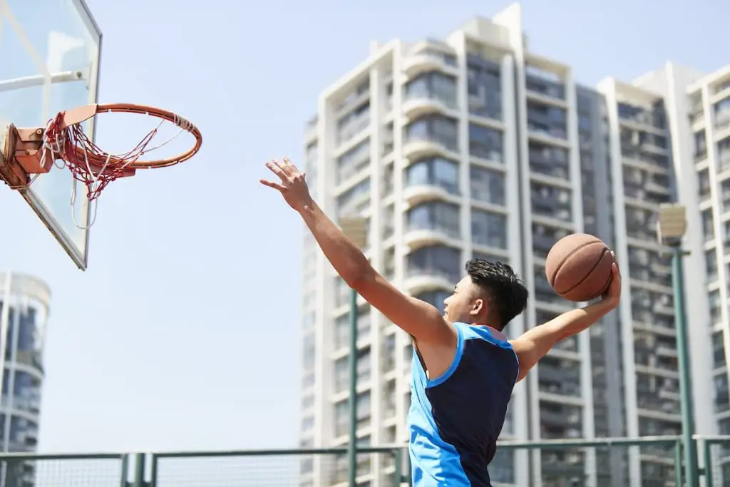 Young asian adult man dunking basketball on outdoor court against city background