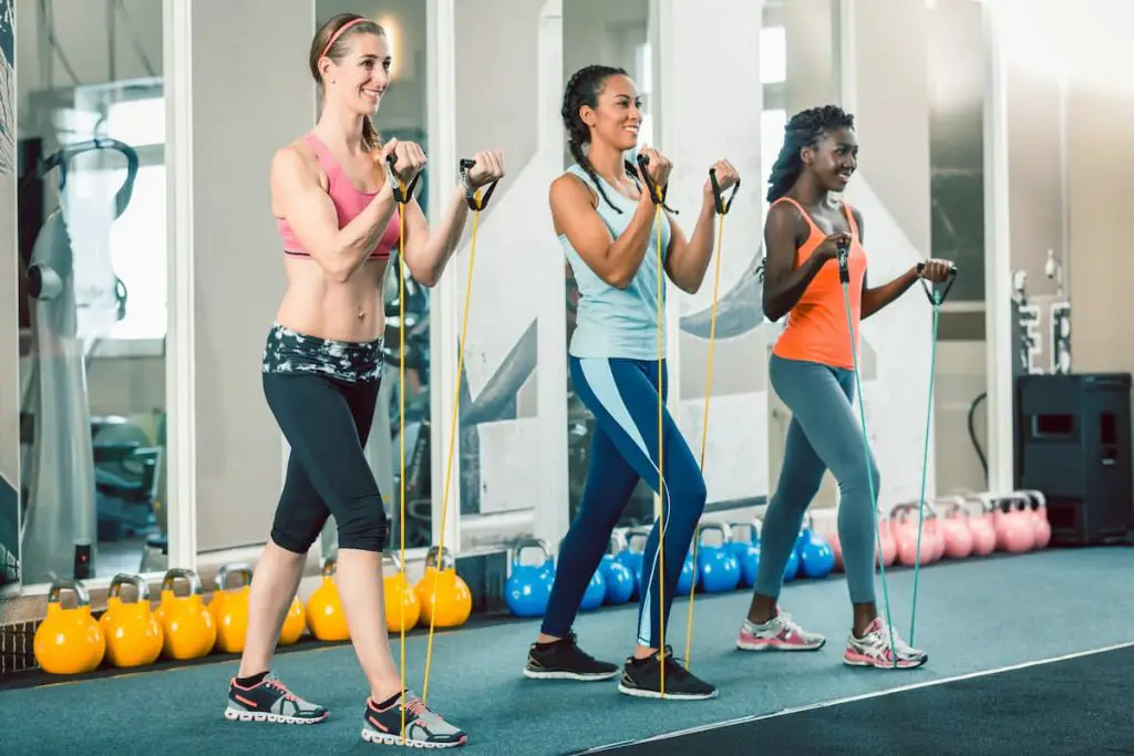 Full length of three fit and beautiful women standing up while exercising with fitness resistance bands during workout class for ladies at the gym